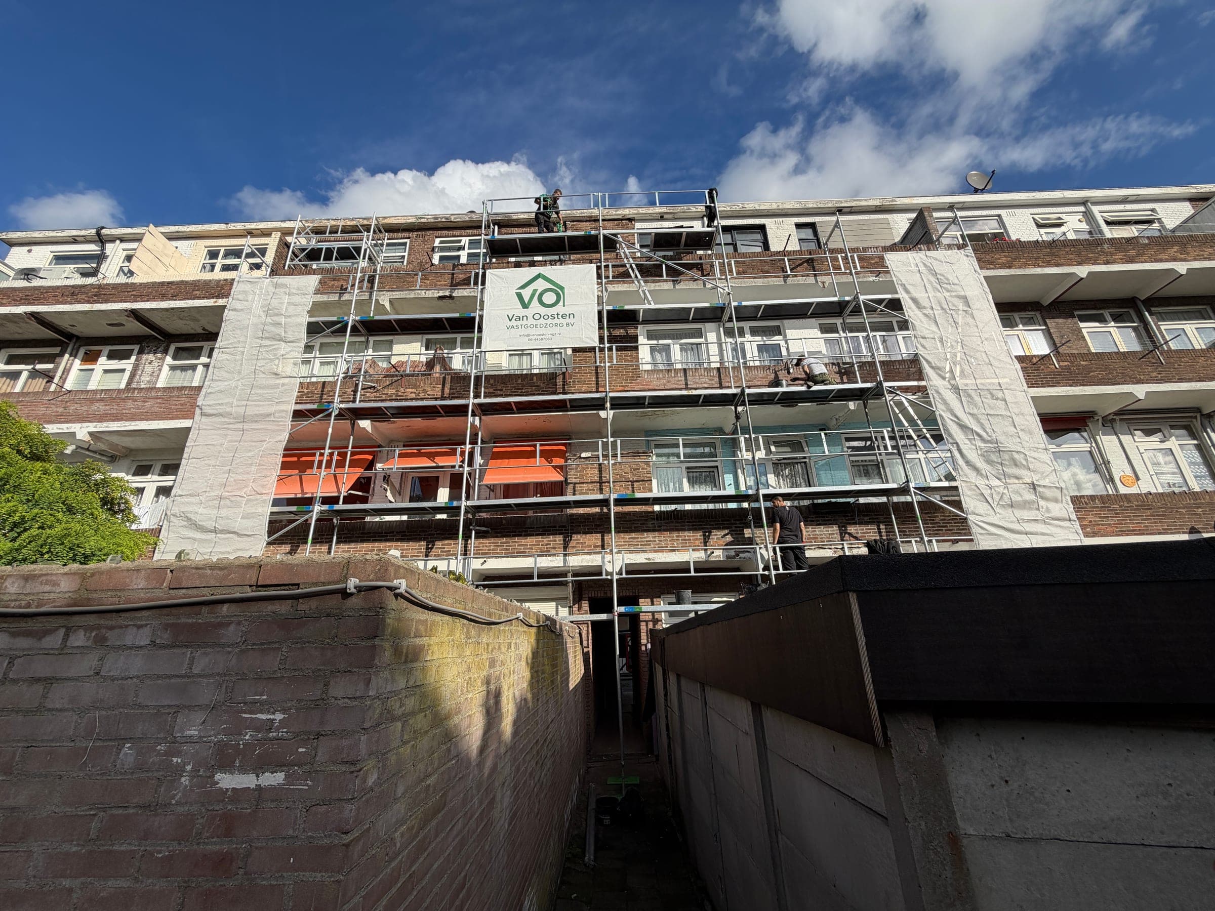 Vakmensen aan het werk aan de gevel en de balkons van een portiekflat, met een spandoek van Van Oosten Vastgoedzorg.